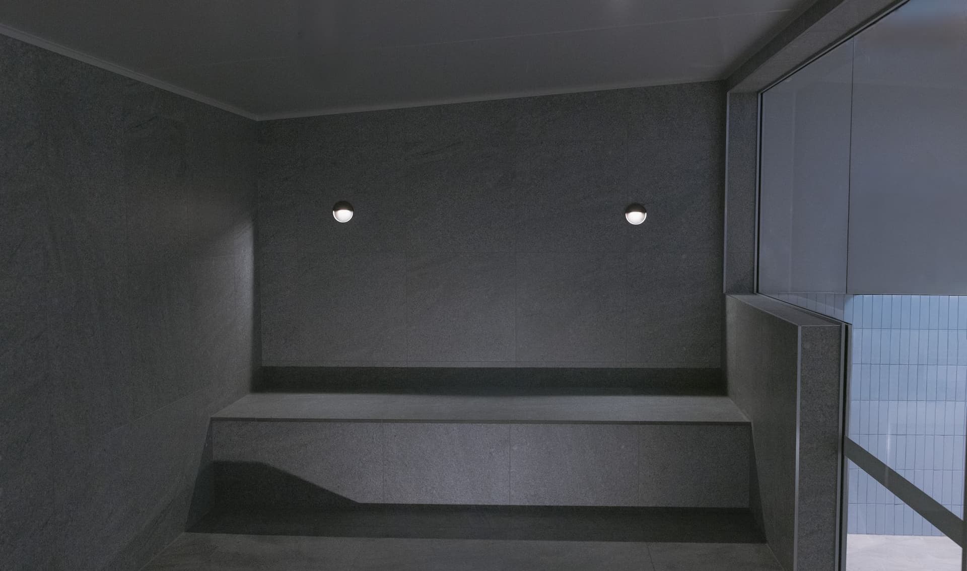 Empty steam room lined in dark grey stone with a long bench, two recessed wall lights and a glazed wall at right