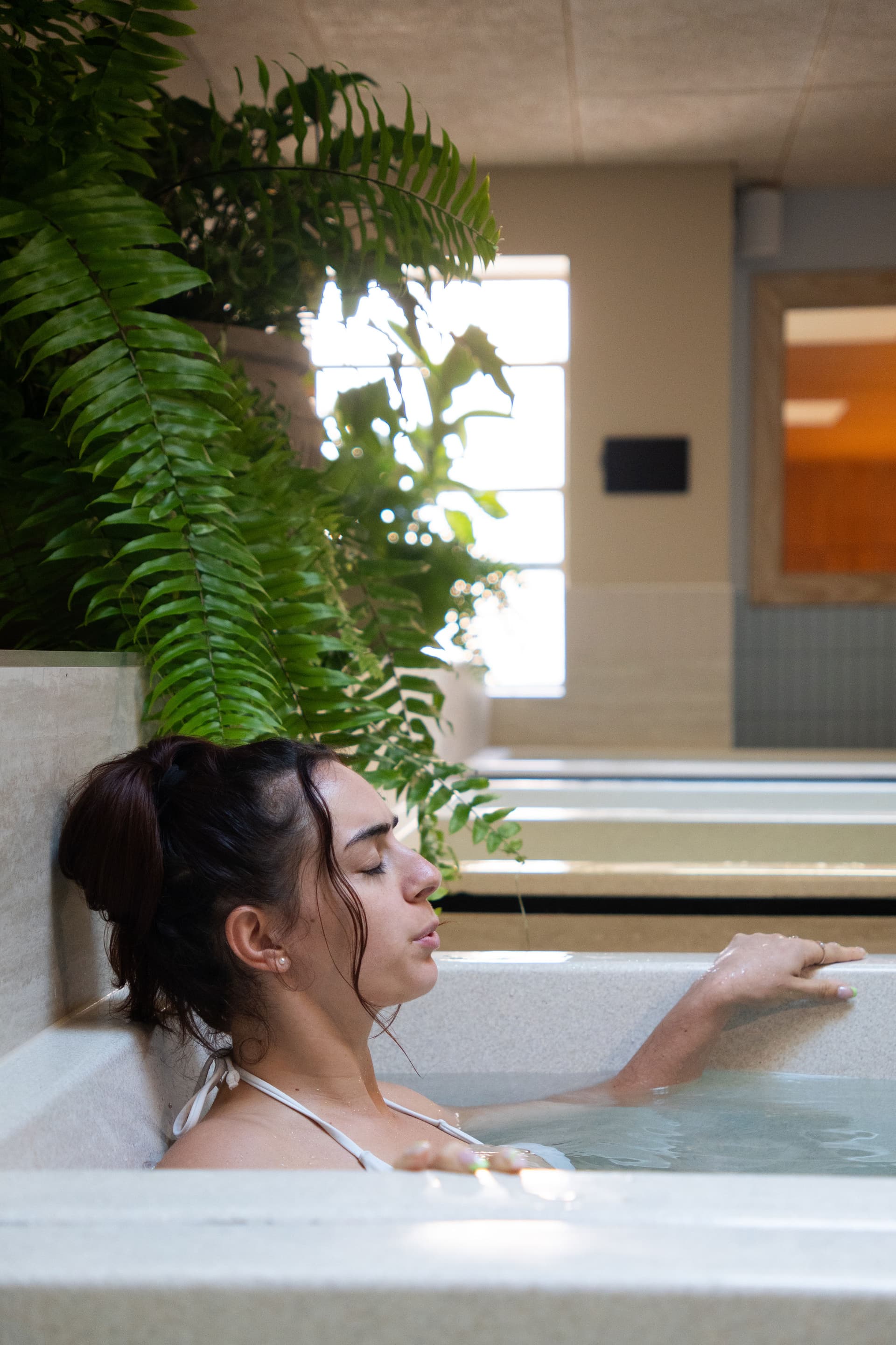 Woman resting an arm on the rim of a cold plunge bath with eyes closed, ferns beside her and a further row of baths behind