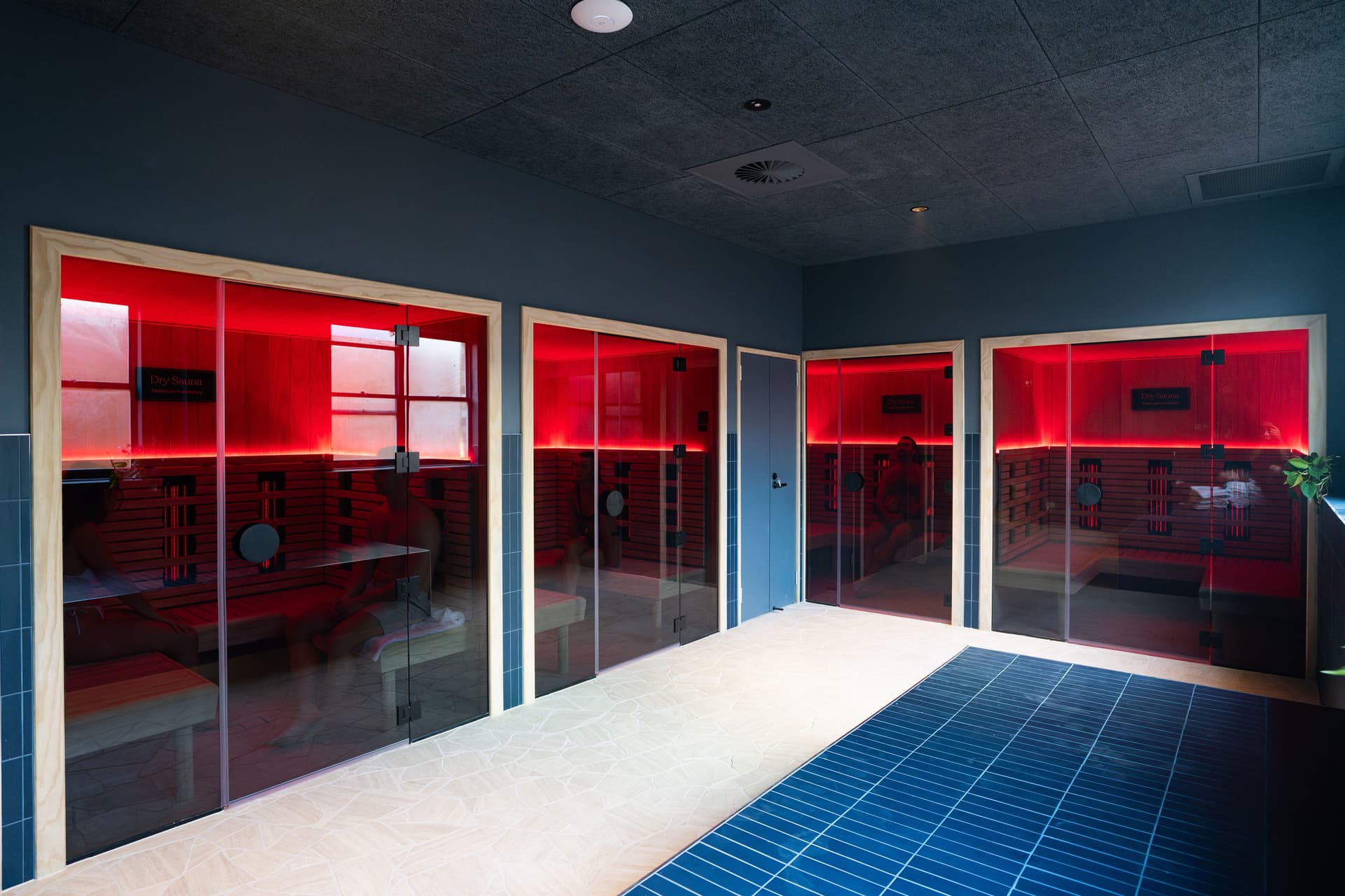 Four infrared sauna cabins behind glass doors, their timber interiors lit red, along a room with a pale stone floor edged by blue tiles