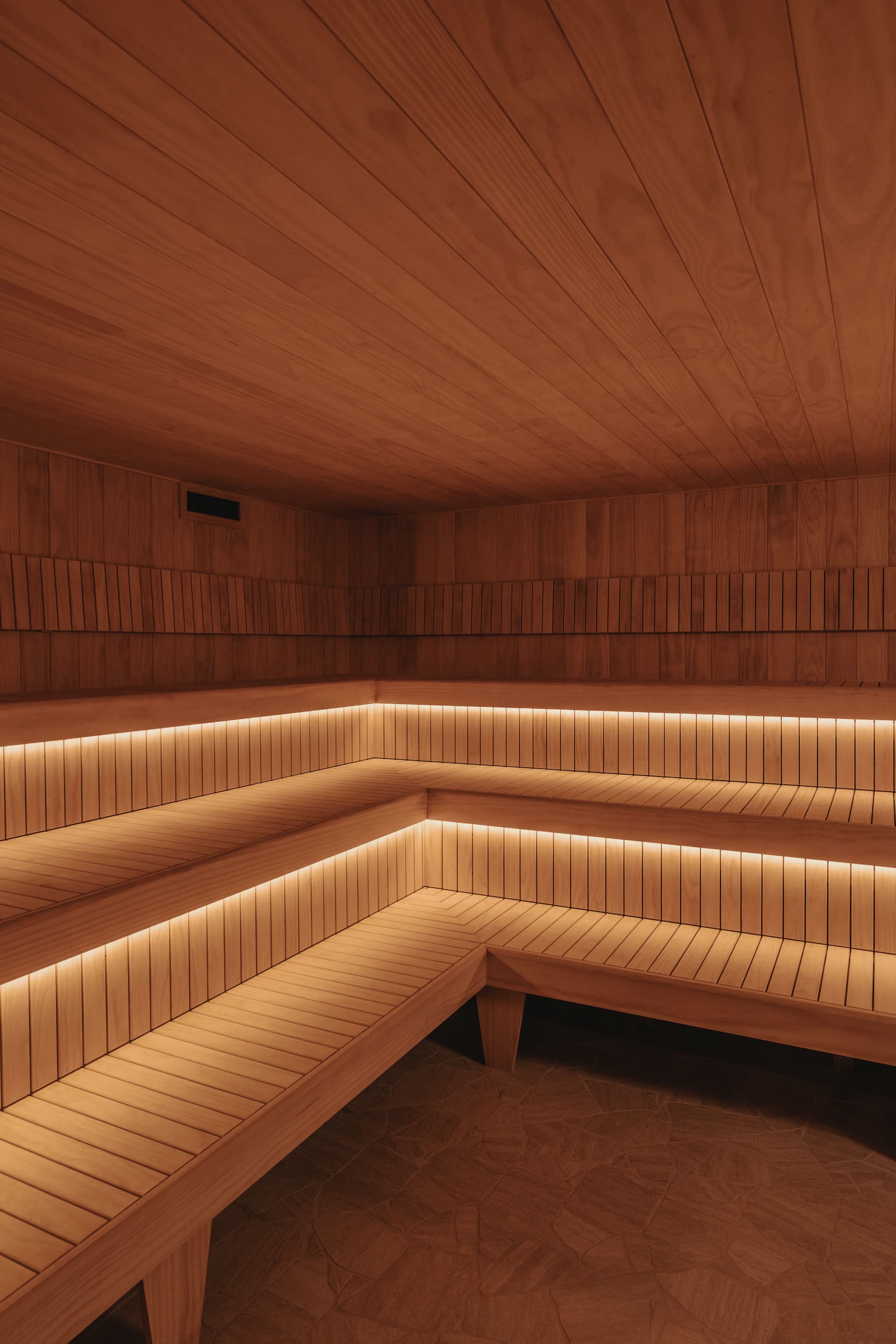 Empty cedar-lined Finnish sauna with two tiers of slatted bench seating wrapping the corner and warm strip lighting beneath each bench