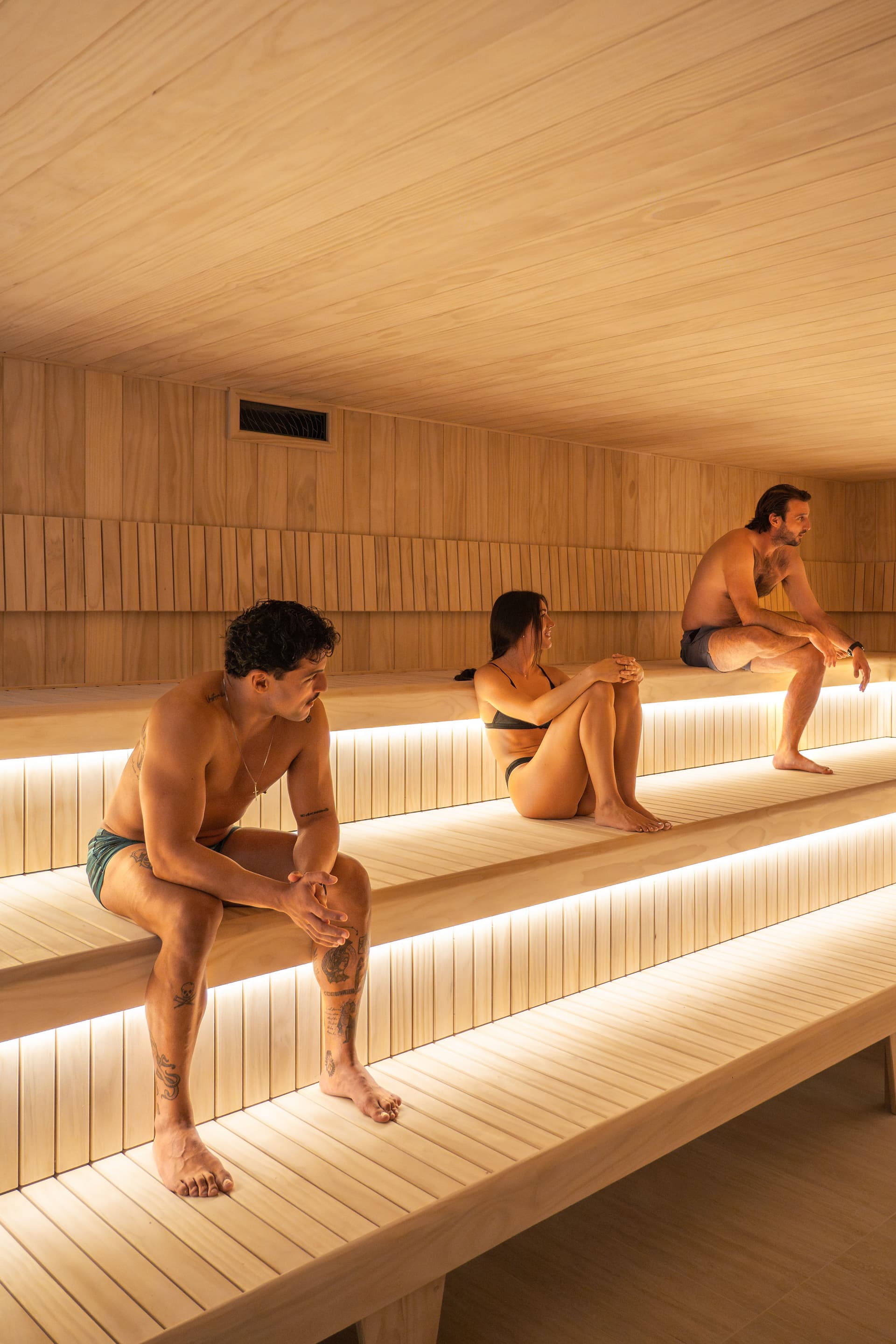 Three people spaced apart on the tiered benches of a cedar-lined Finnish sauna, sitting quietly under warm strip lighting
