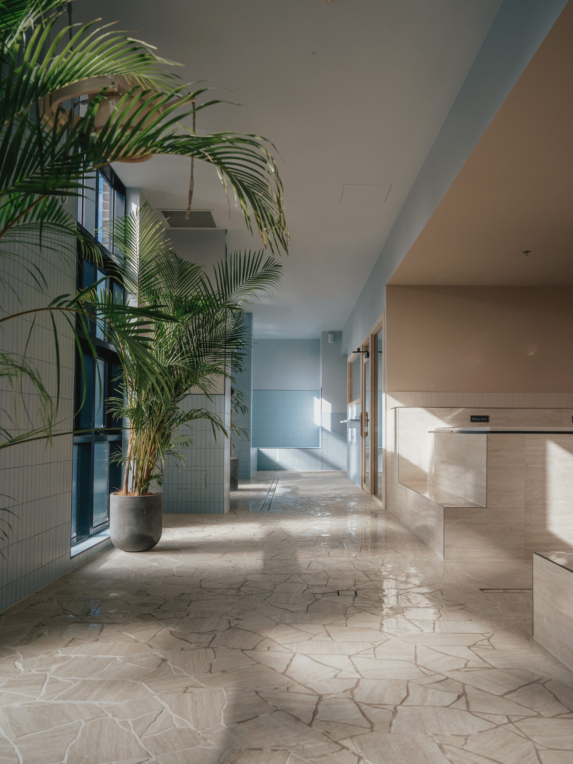 Sunlit walkway with pale tiled walls, a tall potted palm and a polished flagstone floor running past a stone counter towards the bathing rooms